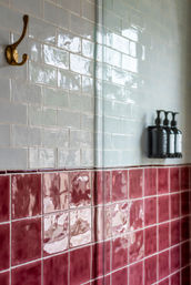 Bathroom shower wall with glossy white subway tiles above ceramic rose-pink square tiles, brass wall hook, glass shower divider and three black wall-mounted pump dispensers.