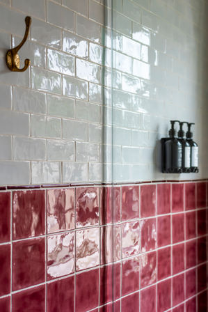 Bathroom shower wall with glossy white subway tiles above ceramic rose-pink square tiles, brass wall hook, glass shower divider and three black wall-mounted pump dispensers.