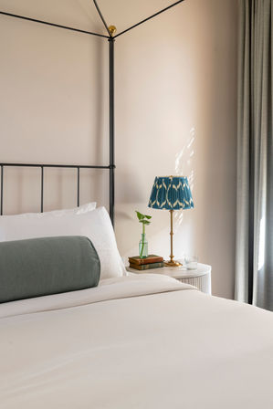 Cozy modern bedroom interior with black iron canopy bed, crisp white linens and gray bolster pillow, round white nightstand holding a brass lamp with blue patterned shade, stacked books, a small green vase and a glass of water, bathed in soft natural light with gray curtains.