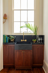 Sunlit kitchen sink nook with black stone farmhouse sink, brass faucet, rich wood cabinets, potted palm, cookbooks and crystal glasses