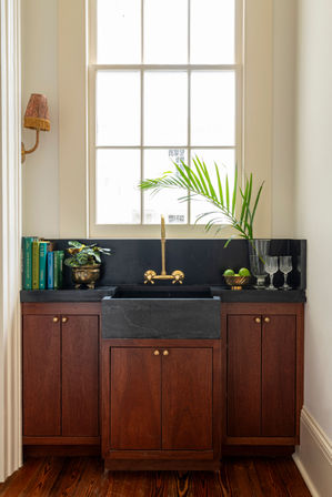 Sunlit kitchen sink nook with black stone farmhouse sink, brass faucet, rich wood cabinets, potted palm, cookbooks and crystal glasses