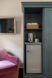 Cozy boutique room corner with teal built-in cabinet holding a retro mini-fridge, espresso machine, mugs and a wicker basket of towels, beside a pink velvet armchair and gold-framed wall TV.