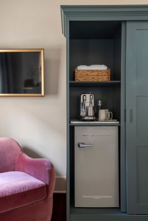 Cozy boutique room corner with teal built-in cabinet holding a retro mini-fridge, espresso machine, mugs and a wicker basket of towels, beside a pink velvet armchair and gold-framed wall TV.