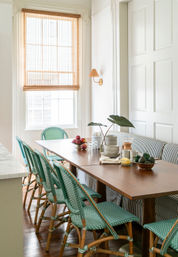 Cheery sunlit kitchen dining nook with a wood table, green woven bistro chairs, striped banquette seating, bamboo window shade, potted plant and bowls of fruit.