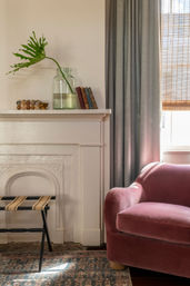 Sunlit cozy living room corner with a blush velvet armchair, white decorative mantel holding a glass vase with a tropical leaf and stacked books, gray curtains, woven blind, and patterned rug.
