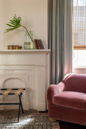Sunlit cozy living room corner with a blush velvet armchair, white decorative mantel holding a glass vase with a tropical leaf and stacked books, gray curtains, woven blind, and patterned rug.