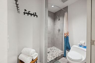 Cozy modern bathroom with pebble-tiled walk-in shower, gray tile walls, curved shower rod with blue beach-themed curtain, white toilet and rolled towels on a stool