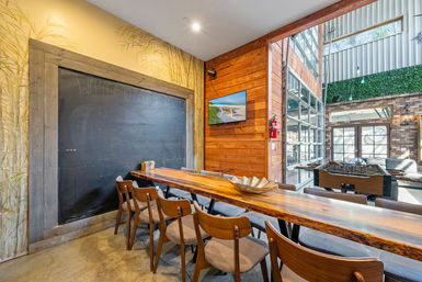 Industrial-chic indoor lounge with a long live-edge communal wood table and mid-century chairs, large chalkboard wall, wood-paneled accent with TV, foosball table and glass garage door opening to a bright seating area.
