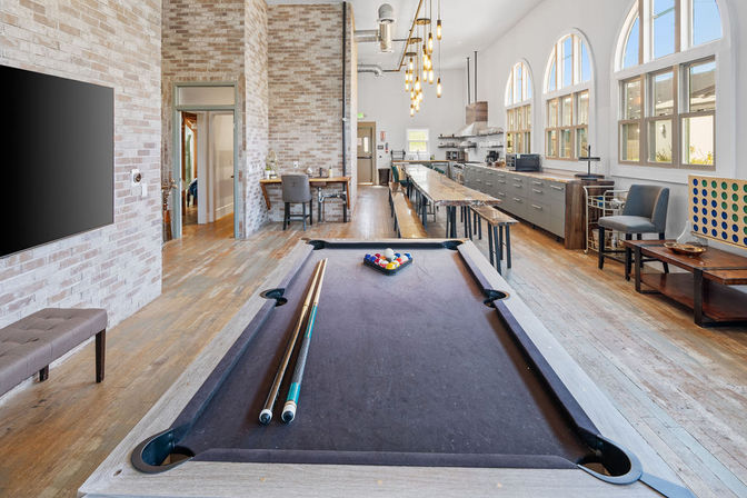 Loft-style communal lounge and game room with a black-felt pool table and cues in the foreground, long wooden communal table, exposed brick walls, industrial pendant lights, arched windows, open kitchen and hardwood floors — playful, modern hangout.