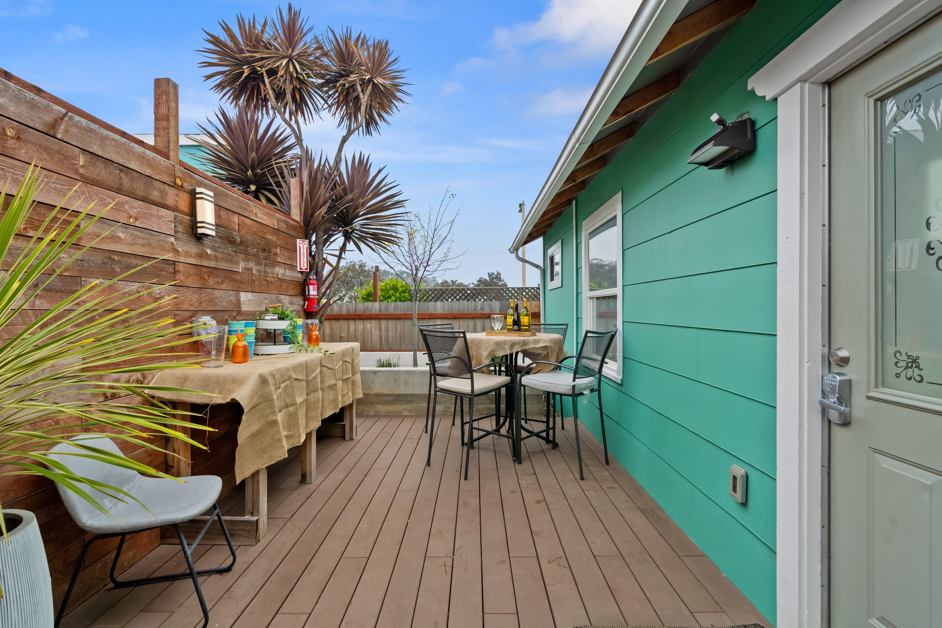 Cozy colorful backyard deck patio with teal house siding, wooden privacy fence, bistro table and chairs, burlap-covered side table and tropical potted plants.