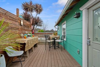 Cozy colorful backyard deck patio with teal house siding, wooden privacy fence, bistro table and chairs, burlap-covered side table and tropical potted plants.