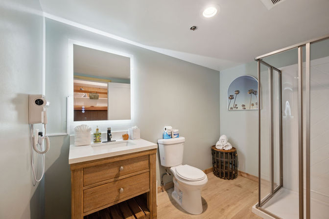 Bright beach-inspired bathroom with wooden vanity and backlit mirror, wall hairdryer, toilet, glass shower, and rolled towels in a wicker basket.