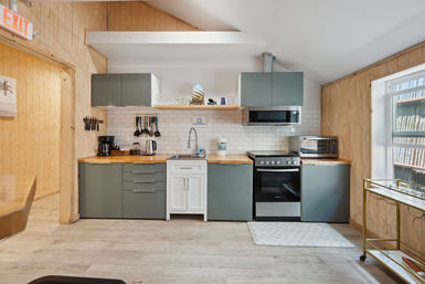 Cozy coastal-style kitchenette with gray cabinets, butcher-block countertops, subway tile backsplash, stainless oven and microwave, coffee maker, white sink and wood-paneled walls.