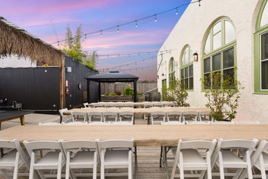 Outdoor courtyard event space with long wooden tables and white folding chairs, string lights overhead, a black gazebo and stucco building with arched green-trim windows under a pink sunset sky
