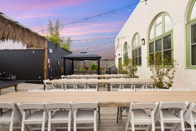 Outdoor courtyard event space with long wooden tables and white folding chairs, string lights overhead, a black gazebo and stucco building with arched green-trim windows under a pink sunset sky