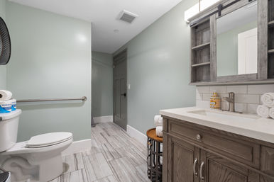 Bright modern guest bathroom with gray-green walls, wood vanity and mirrored cabinet, white quartz countertop, subway tile backsplash, chrome faucet, rolled towels, toilet with safety grab bar, and gray wood-look floor tile — spa-like style.