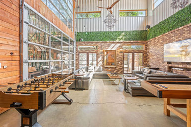 Bright modern industrial game lounge with foosball and shuffleboard tables, oversized leather sectional seating, exposed brick walls, floor-to-ceiling glass garage door and green wall accents.