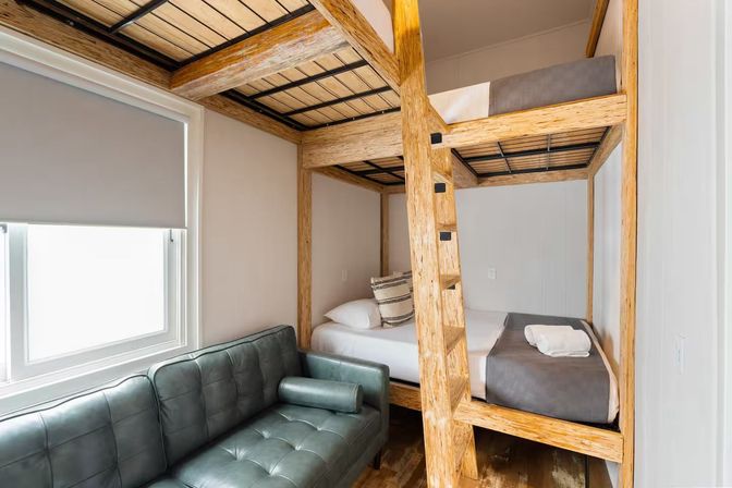 Cozy modern bunk room with rustic wooden double bunk bed and ladder, gray linens with folded towel, tufted green leather sofa beside a window with roller shade.