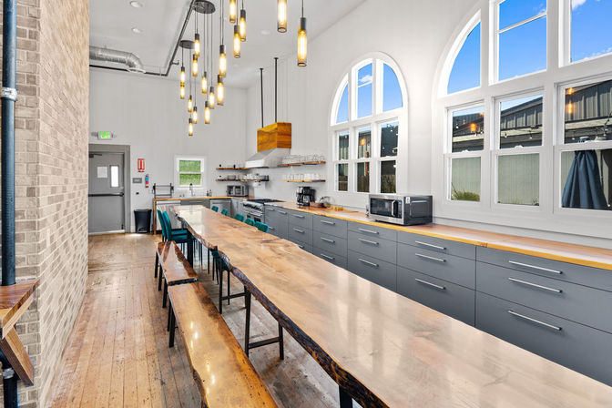 Bright industrial-style communal kitchen in a renovated loft with a long live-edge wood table and bench seating, hanging pendant lights, arched windows, gray lower cabinets, wood floors and stainless appliances.