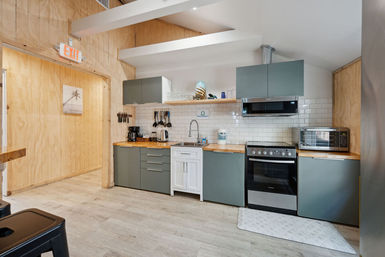 Bright modern kitchenette with gray cabinets, wood countertops, white subway tile backsplash, stainless stove and microwave, coffee maker, hanging utensils and exposed ceiling beams.
