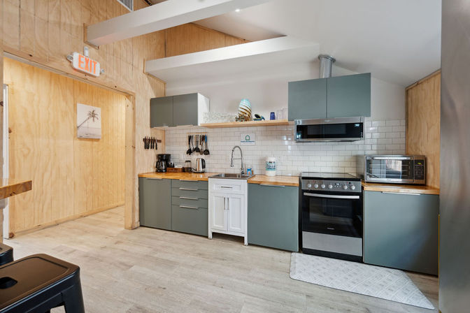 Bright modern kitchenette with gray cabinets, wood countertops, white subway tile backsplash, stainless stove and microwave, coffee maker, hanging utensils and exposed ceiling beams.