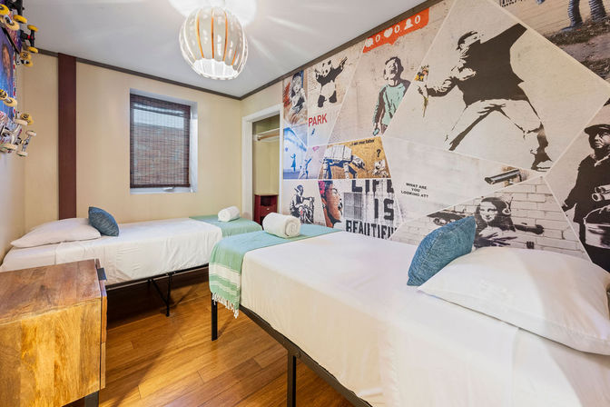 Bright urban-style twin bedroom with two single beds in white linens, blue pillows and green throws, hardwood floor, modern pendant light, bamboo-blind window and large graffiti-inspired mural wall art