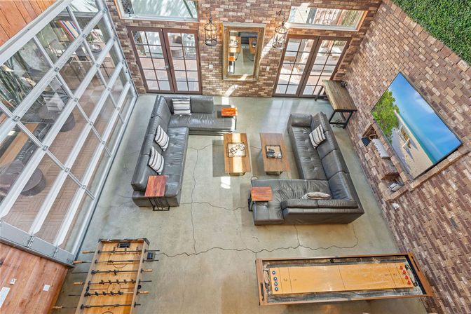 Sunlit, game-ready urban loft living area with exposed brick walls and concrete floor, a large glass garage-door wall, two dark leather sectional sofas, wall-mounted TV, coffee tables, foosball and shuffleboard.