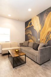 Modern living room with a gray sofa and patterned loveseat, rustic wood-and-metal coffee table with a gold bowl, and a bold dark accent wall featuring oversized gold leaf mural under recessed lighting.