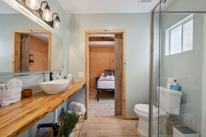 Bright modern rustic bathroom with live-edge wooden vanity and vessel sink, glass-enclosed shower, toilet, and doorway opening to a cozy wood-accent bedroom