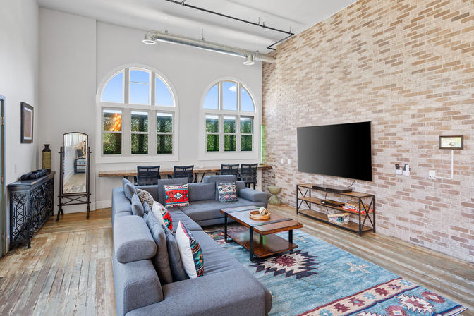 Bright modern loft living room with exposed light brick wall, arched windows and industrial ductwork, gray sectional sofa with colorful southwestern pillows, wooden coffee table, bohemian rug, wall-mounted flat-screen TV and a window desk with chairs on hardwood floors.