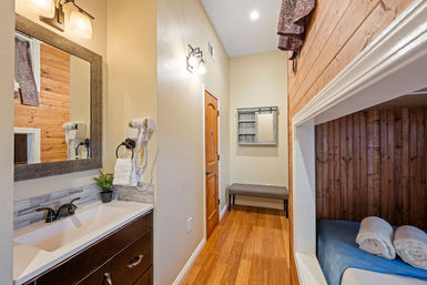 Cozy rustic cabin interior with bathroom vanity and mirror, wall-mounted hair dryer, hardwood hallway with bench and mirror, and a wood-paneled sleeping nook with rolled towels