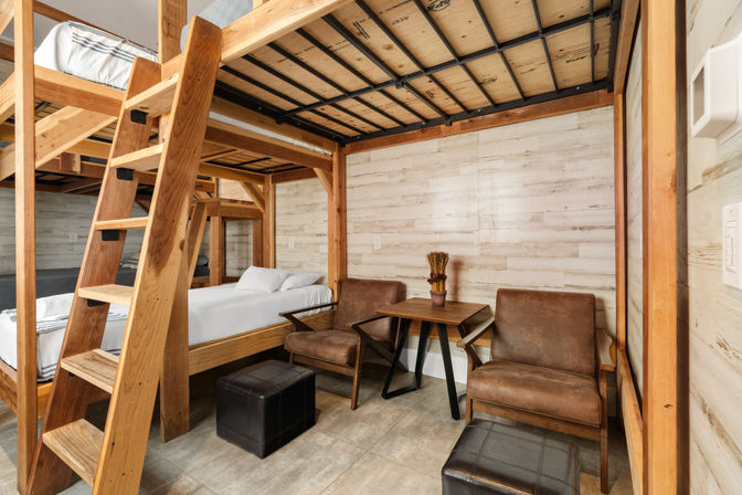 Cozy cabin-style interior with sturdy wooden bunk beds and ladder, twin beds, light wood plank walls, two brown leather armchairs, small wooden table and leather ottomans.