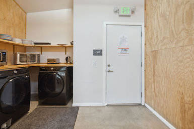 Bright communal laundry room with two black front‑load washers under a wooden counter, microwave, toaster oven and kettle on open shelves, and a white exit door with a green EXIT sign above.