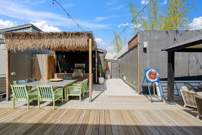 Sunny urban rooftop patio with wooden deck, thatched pergola over a large dining table with mint-green chairs, built-in grill, string lights and a colorful cornhole target