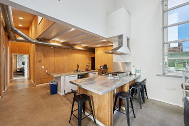 Modern industrial loft kitchen with marble-look peninsula and three black metal bar stools, stainless steel range hood, plywood walls, exposed ductwork, concrete floor and garage-style glass window.