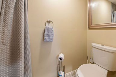 Compact bathroom interior with white toilet, freestanding toilet-paper holder, light blue hand towel on a wall ring, framed mirror, beige walls and a textured gray shower curtain.