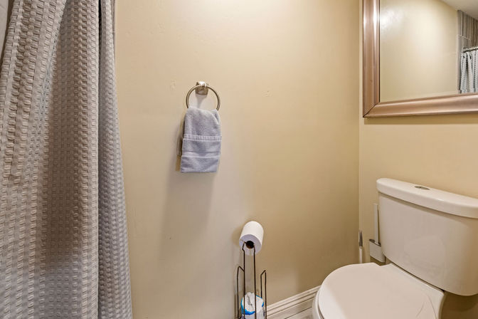 Compact bathroom interior with white toilet, freestanding toilet-paper holder, light blue hand towel on a wall ring, framed mirror, beige walls and a textured gray shower curtain.