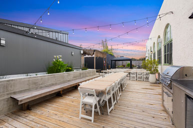 Sunset-lit outdoor wooden deck patio with long banquet tables, white folding chairs, string lights, pergola and stainless grill