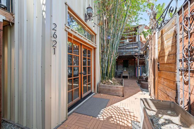 Urban narrow backyard patio with corrugated metal siding and wooden French doors, tall bamboo in planters, string lights overhead, paver deck tiles, decorative wooden fence with ironwork and a cozy covered outdoor seating area
