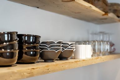 Rustic wooden kitchen shelf lined with stacked dark ceramic bowls, scalloped gray bowls with white rims, a white mug and clear glassware in soft focus