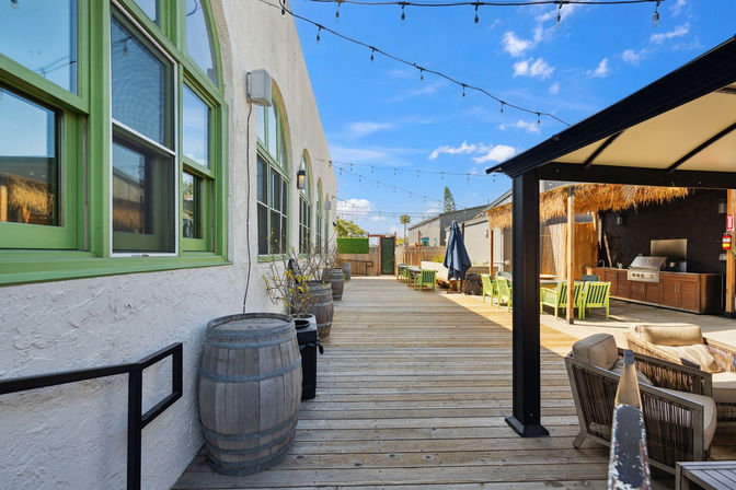 Sunny outdoor wooden-deck patio with green-trim windows, wine barrels, string lights, shaded lounge area and outdoor grill seating under thatch