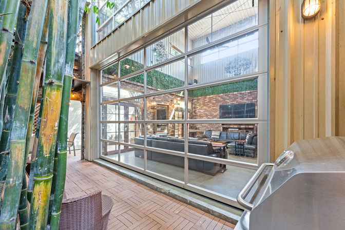 Loft-style indoor-outdoor living space with bamboo-lined patio, stainless grill, large glass roll-up garage door opening to a cozy lounge with leather sofas and exposed brick TV wall.