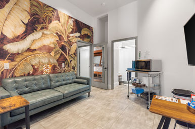 Modern urban lounge with teal leather sofa, oversized tropical wall mural, tiled floor, microwave cart and doorway revealing a bunk-bed room.
