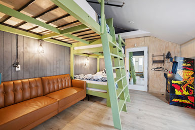 Bright playful bedroom with lime-green loft bunk bed and ladder, lower double bed with blue patterned bedding, caramel tufted leather sofa against gray wood-paneled wall, industrial hanging lights, retro arcade cabinet and light wood floors.