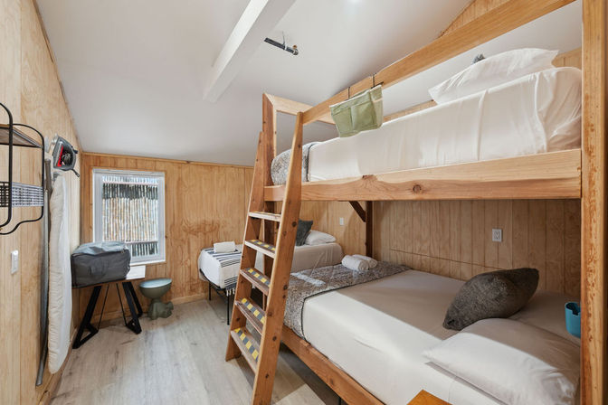 Wood-paneled cabin bedroom with laddered wooden bunk bed, extra single bed by a window, light laminate floor and neutral linens in a compact, cozy guest room.