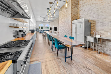 Bright industrial loft communal kitchen and dining area with a long live-edge wood table, teal chairs, stainless steel appliances, exposed brick, pendant lights and arched windows.