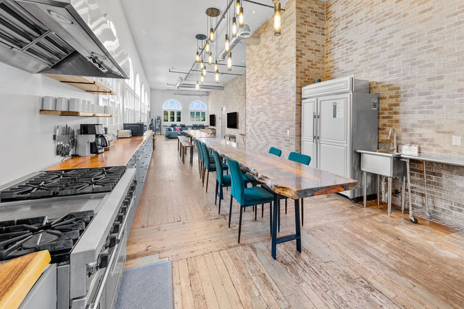 Bright industrial loft communal kitchen and dining area with a long live-edge wood table, teal chairs, stainless steel appliances, exposed brick, pendant lights and arched windows.