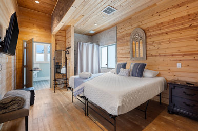 Cozy wood-paneled cabin bedroom with vaulted pine ceiling, king bed dressed in white quilt and blue accent pillows, wall-mounted TV, full-length mirror, bench, and an en-suite bathroom with patterned tile visible