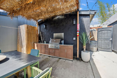 Outdoor patio kitchen with stainless steel grill and wood cabinets under a tropical thatch pergola, concrete patio, bamboo privacy screen, dining table and potted yucca
