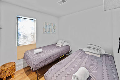 Cozy guest room with two twin beds dressed in gray geometric quilts, rolled towels and striped pillows, wood floor, small window and abstract wall art.